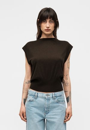 Moteris su tamsiais plaukais, dėvinti juodą rankovių neturintį viršutinį drabužį ir šviesiai mėlynas džinsines kelnės, rodanti tatuiruotes ant abiejų rankų prieš paprastą foną.