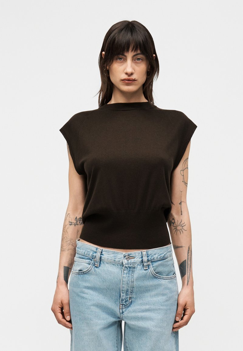 Femme aux cheveux foncés portant un haut noir sans manches et un jean bleu clair, montrant des tatouages sur les deux bras sur un fond uni.