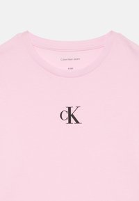 Világos rózsaszín Calvin Klein Jeans póló fekete "cK" logóval, ami középen található a kerek nyak körül, 8-as méret (128) látható a gallér belsejében.