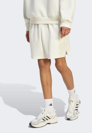 Osoba ubrana w kremowe szorty i pasującą bluzę, białe buty Adidas i białe skarpetki Adidas, stojąca na jasnym tle.