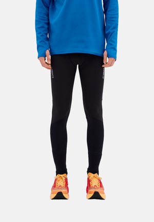 Person wearing black athletic leggings, blue long-sleeve top, and red-orange running shoes, standing straight against a plain background.