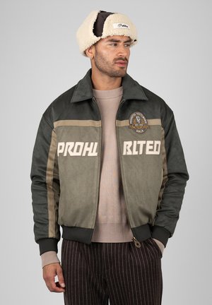 Mand iført grøn og beige jakke med tekst og logo, stribede mørke bukser, en beige sweater og en beige shearling fælderhat, der kigger til siden.