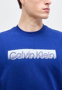 Ung person iført en kongeblå sweatshirt med et hvidt rektangulært logo, der viser "Calvin Klein" i lyse lilla bogstaver.