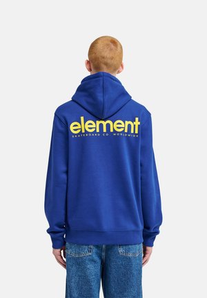 Osoba z krótkimi rudymi włosami nosząca niebieską bluzę z kapturem z żółtym tekstem "element skateboard co. worldwide" na plecach oraz niebieskie dżinsy.