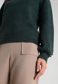 Vihreä neulepaita, jossa on ribattu helma ja helmassa helmiä, yhdistettynä beigeihin räätälöityihin housuihin, joissa on sivutaskut ja ompeluominaisuuksia.