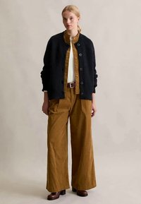 Kvinna iförd en svart knäppt kofta över en brun knäppt skjorta, med vida bruna byxor och bruna klackskor, stående mot en neutral bakgrund.