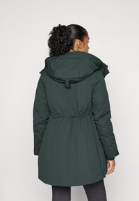 Veste rembourrée vert foncé avec taille à cordon de serrage, grande capuche et col montant. Comprend des manches longues et une texture lisse et mate.