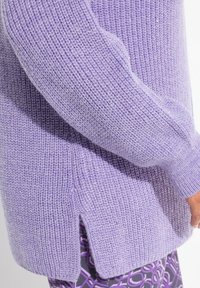 Lila stickad tröja med ribbat mönster, sidoklyftor och glittrande detaljer. Har en avslappnad passform och lätt puffade ärmar.