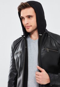 Chaqueta de cuero negra con capucha, con bolsillos con cremallera y detalles acolchados en los hombros, llevada sobre una camisa gris claro.