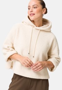 Haut à capuche en tissu doux couleur crème avec une texture en polaire, grande capuche, cordon de serrage avec des embouts métalliques, et manches larges. Porté avec des pantalons marron.
