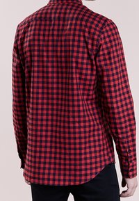 Hombre con una camisa de cuadros roja y negra de manga larga, visto desde atrás, combinado con pantalones negros.