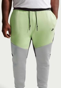 Homme portant un pantalon de survêtement Nike vert clair et gris avec des cordons noirs et un logo Nike noir sur la cuisse, les mains dans les poches.