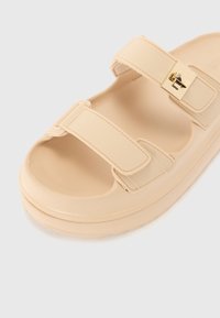 Beige sandál s dvěma nastavovacími popruhy a zlatým přezkou. Má polstrovanou podrážku a hladký povrch.