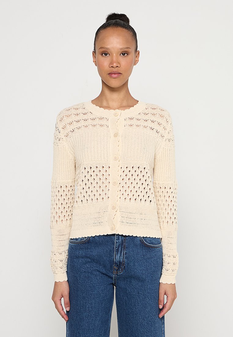 Kobieta z włosami upiętymi w kok, ubrana w kremowy sweter zapinany na guziki z wzorzystymi dziurkami oraz niebieskie dżinsy, stojąca na jednolitym tle.