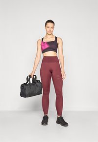 Sportbeha in zwart met roze tie-dye, hooggewaist bordeauxrode leggings en zwarte sneakers. Houdt een zwarte sporttas vast.