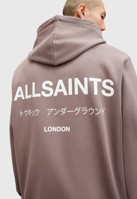 Hættetrøje i taupe med en blød tekstur, der har fed hvid tekst. Teksten inkluderer "ALLSAINTS," japanske tegn, og "LONDON."