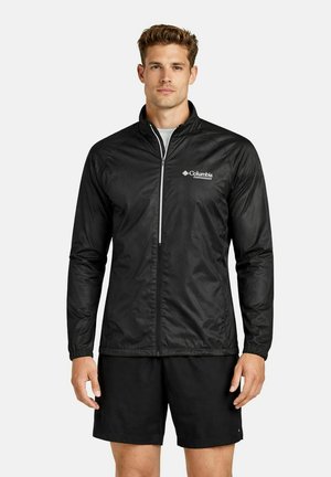 Giovane uomo con giacca Columbia nera con zip completa e pantaloncini neri, in piedi davanti a uno sfondo bianco uniforme.