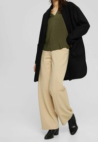 Manteau noir surdimensionné, haut plissé vert olive, pantalon ample beige et bottines à talons noires. Textures lisses et couleurs contrastées.