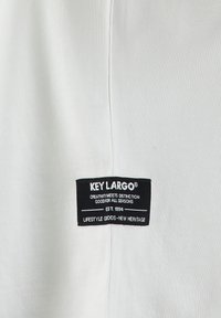 Tela blanca con textura acanalada que presenta una etiqueta negra. La etiqueta muestra "KEY LARGO" y texto adicional en detalles blancos y rojos.