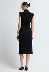Czarna sukienka bez rękawów z wysokim kołnierzem, paskiem w talii, dopasowaną sylwetką i długością do kolan, sparowana z czarnymi szpilkami. Gładka faktura materiału.