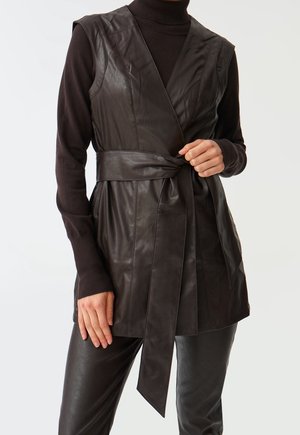 Personne portant un gilet sans manches en cuir noir noué à la taille sur un col roulé noir à manches longues avec un pantalon en cuir noir assorti.