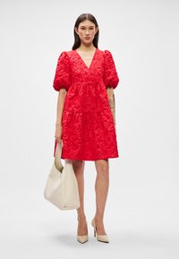 Raudona su pūstomis rankovėmis suknelė su sluoksniuotu dizainu, tekstūruotu audiniu ir giliu V formos kaklu. Pora su smėlio spalvos rankine ir kūno spalvos aukštakulniais.