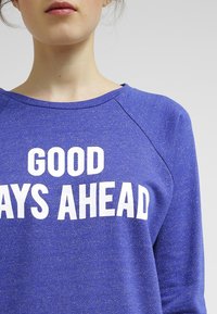 Sudadera azul real con una tela texturizada. Presenta un escote redondo y un texto blanco que dice "BUENOS DÍAS POR DELANTE" en la parte frontal.