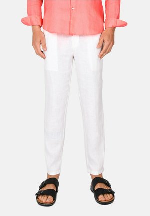 Uomo che indossa una camicia corallo con bottoni, pantaloni di lino bianchi e sandali neri con fibbia, in piedi contro uno sfondo semplice.