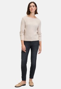 Hellbeiger, gerippter Pullover mit weitem Ausschnitt, langen Ärmeln und eng anliegenden Bündchen, kombiniert mit dunklen Jeans und beigen Slippern.