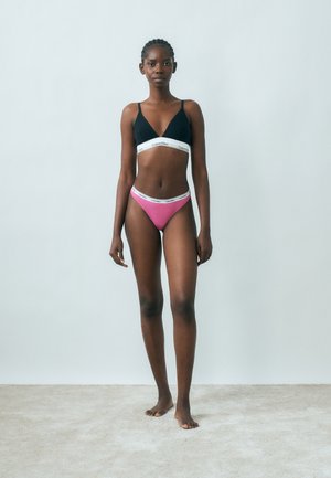 Mladá žena stojaca naboso opretá o obyčajnú sivú stenu, oblečená v čiernom a ružovom spodnom prádle značky Calvin Klein.