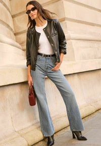 Veste en cuir synthétique noire à boutonnières, chemise blanche ajustée, jeans à jambe large bleu clair, bottines à talons noires et un petit sac à main bordeaux.