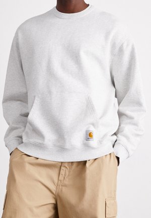 Mand iført lys grå Carhartt sweatshirt med frontlomme og khaki cargobukser, stående med hænderne i lommerne mod en hvid baggrund.