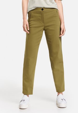 Pantalons ajustés vert olive avec une coupe slim, poches zippées et fermeture à bouton, portés avec des baskets blanches à plateforme. Tissu lisse et texturé.