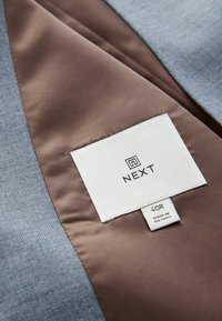 Interiorul unui jachet textil gri, căptușit cu satin maro. Are o etichetă albă cu „NEXT”, mărimea „40R” și „FABRICAT ÎN VIETNAM”.