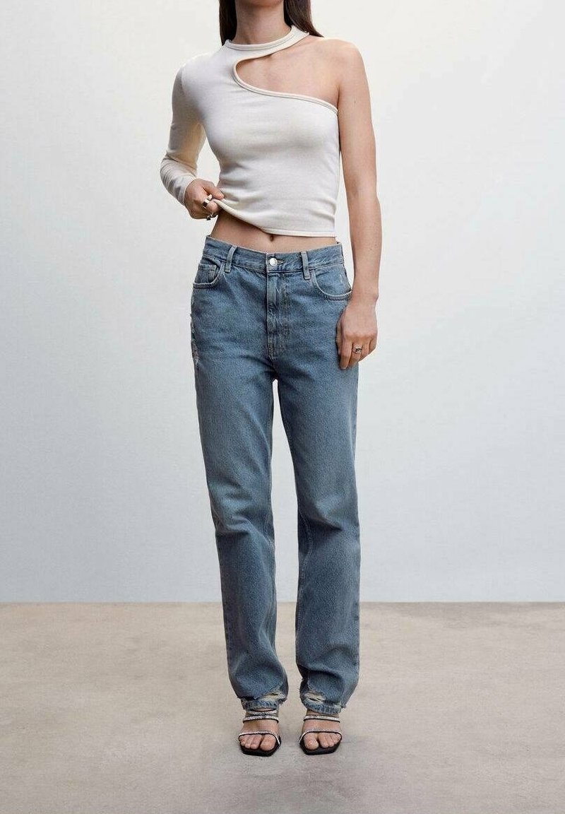 Femme portant un haut blanc asymétrique à une manche, un jean bleu avec des déchirures aux ourlets, et des sandales noires à brides, debout contre un mur uni.