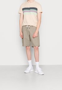 Persoon in beige shorts met witte trekkoord, beige t-shirt met pastel horizontale strepen, witte sokken en witte sneakers, staand op een grijze vloer.