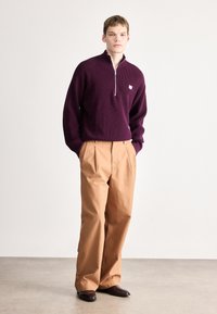 Bordó félcipzáros pulóver bézs nadrággal; a pulóver texturált felületű és kis logóval díszített.