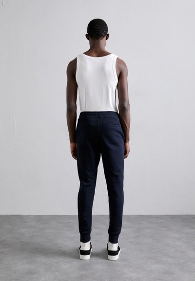 Αρσενικός που στέκεται με την πλάτη προς την κάμερα, φοράει άσπρο φανελάκι, ναυτικό παντελόνι φόρμας, άσπρες κάλτσες και μαύρα αθλητικά παπούτσια σε γκρίζο δάπεδο και απλό τοίχο.