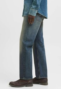Jeans din denim de culoare albastră închisă, cu croială dreaptă, detalii ușor decolorate și cusături, asortați cu pantofi bărbătești cu șireturi de culoare maro.