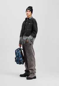 Sort og grå denimskjorte, løstsiddende grå jeans, sort beanie og en blå camouflagemønsteret rygsæk med justerbare stropper. Sorte fodtøj.