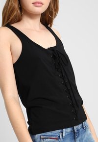 Nainen, jolla on päällä musta hihaton paita, jossa on edessä nyöritys, ja siniset farkut, sekä suuret renkaat korvissa.