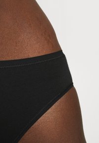 Schwarzes Baumwoll-Bikini-Unterwäsche mit glatter Textur, elastischem Bund und flachen Nähten, teilweise vor dunklerer Haut gezeigt.