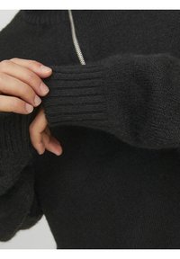 Czarny dziergany sweter z prążkowanymi mankietami i srebrnym zamkiem błyskawicznym. Tkanina jest miękka i teksturowana, na zdjęciu zbliżenie na rękaw i detal dłoni.