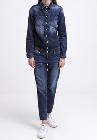 Chaqueta de mezclilla con dos bolsillos en el pecho y botones plateados, color azul oscuro deslavado, combinada con jeans a juego y zapatillas blancas.