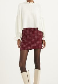 Blusa bianca corta con scollatura arricciata e lunghe maniche a sbuffo, abbinata a una minigonna a quadri rossi e stivali beige al ginocchio con zip.