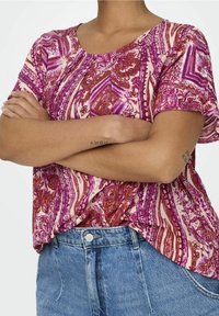 Pink and purple patterned top with a round neckline, short sleeves, and a relaxed fit, paired with blue high-waisted jeans.