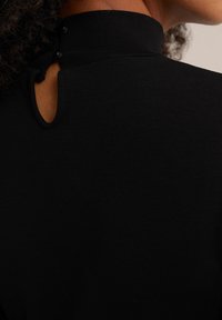 Schwarzes Oberteil mit hohem Kragen, das eine Schlüsselöffnungsöffnung hinten mit zwei runden Knöpfen und einer glatten, dehnbaren Stofftextur aufweist.