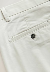 Poche arrière d’un pantalon beige clair avec un seul bouton foncé, passants visibles et couture détaillée sur tissu lisse.