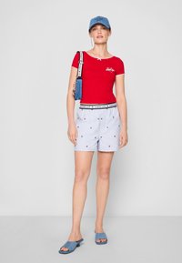 Rød t-skjorte med korte ermer, hvit kant og logo, kombinert med lyseblå stripete shorts med stjernetern, og blå slidesandaler.