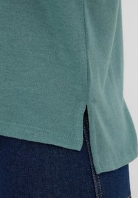 Top a maglione color teal con una piccola apertura laterale, tessuto leggermente testurizzato e orlo dritto, abbinato a pantaloni in denim scuro.